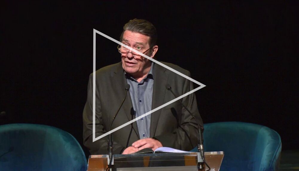 The actor Ciarán Hinds stands behind a lecturn on a darkened Royal Festival Hall stage as he reads from Seamus Heaney: A Life in Letters. Hinds is wearing a tweed jacket and glasses; his dark hair is combed back.