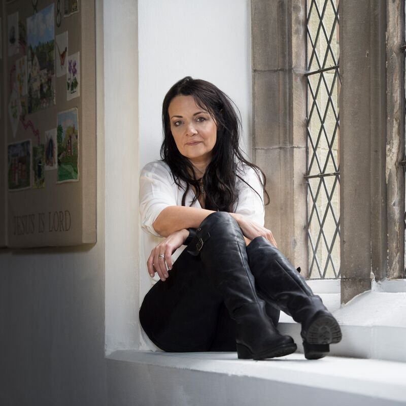 Author Joanna Cannon sits beside a window. She wears a white top, black trousers and black boots