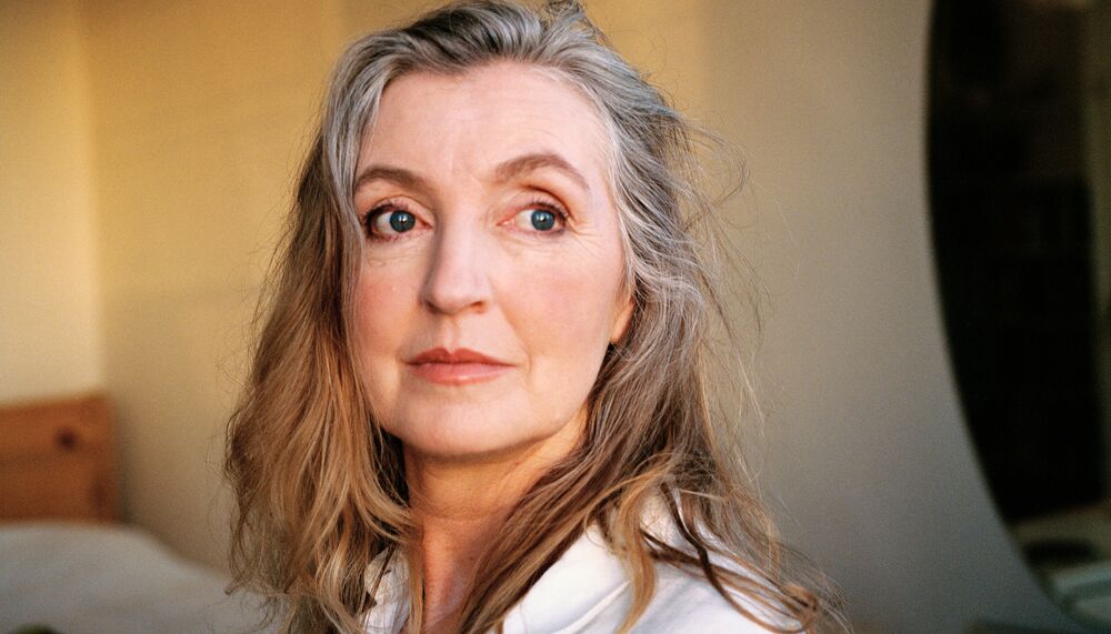 Rebecca Solnit wears a white shirt and is pictured at a 3/4 angle in a dusk lit room. Her dark grey/brown hair is swept back off her face.
