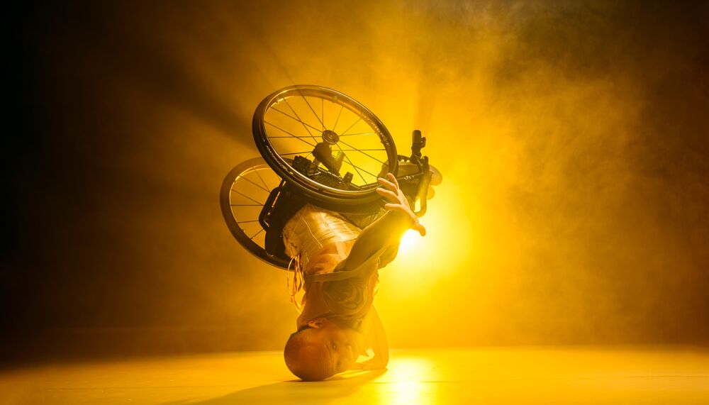 A dancer in a wheelchair leans upside down on the head with the wheelchair in the air in front of a bright yellow light.