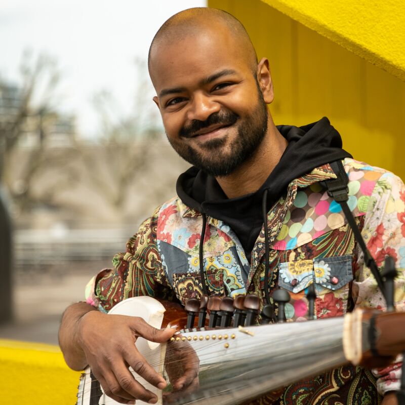 Soumik Datta smiling and holding his sarod in front a yellow staricase