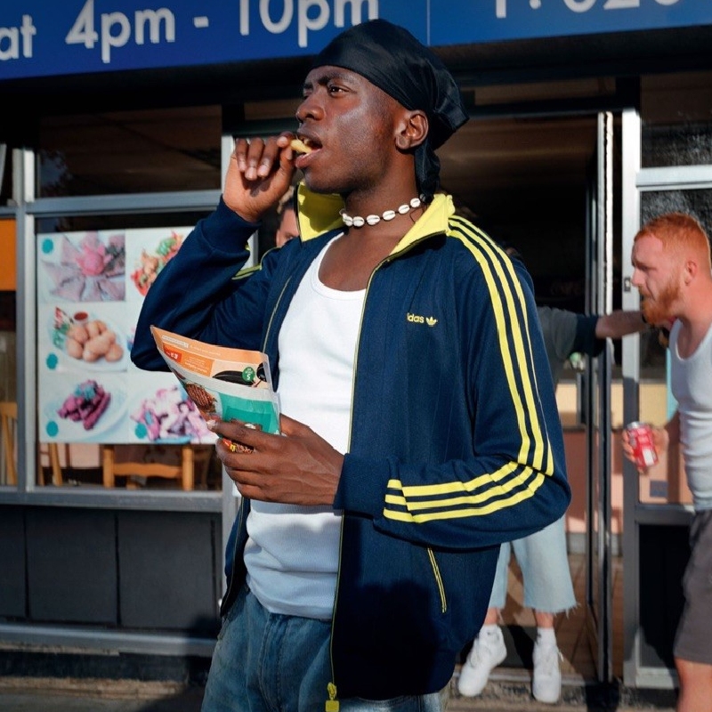 Musician Master Peace standing outside of a shop holding a paper cone of chips and putting one chip into his mouth. He wears a navy blue jacket with yellow stripes and a black do-rag on his head.