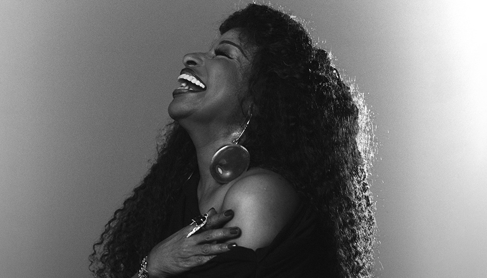 Black and white side profile of the singer Chaka Khan, laughing. She is wearing an over the shoulder black dress.