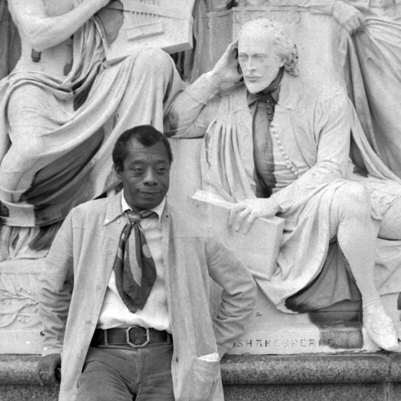 Black and white photo of author James Baldwin standing in front of a statue