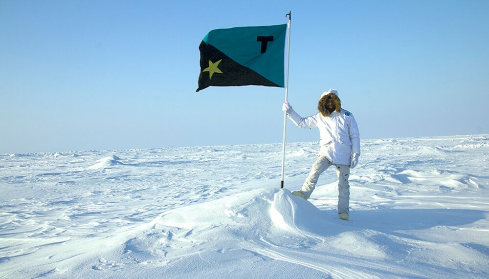 Artist Tavares Strachan, a Black man in his 30s, stands at the North Pole on a white landscape of ice and snow, wearing an all white snowsuit. He is holding a flag pole, on top of which is a flag that is aquamarine and black divided diagonally with a Gold star on the lower black half and a black 't' on the upper aquamarine half