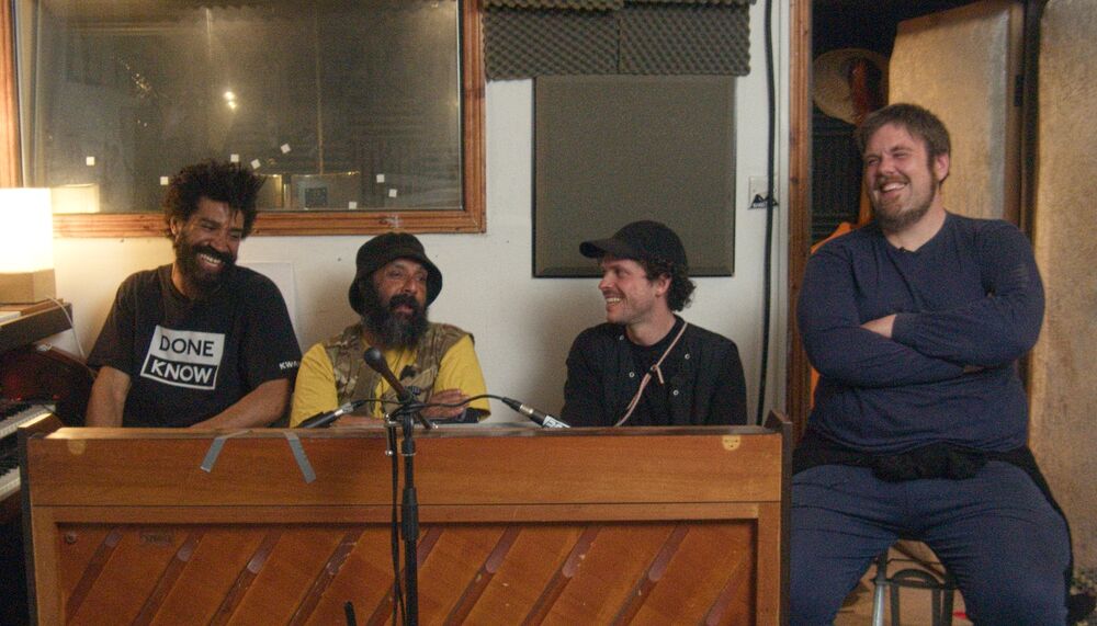 The four members of Speakers Corner Quartet share a joke as they sit together in a music studio; one sits on a stool, the other three behind an upright piano