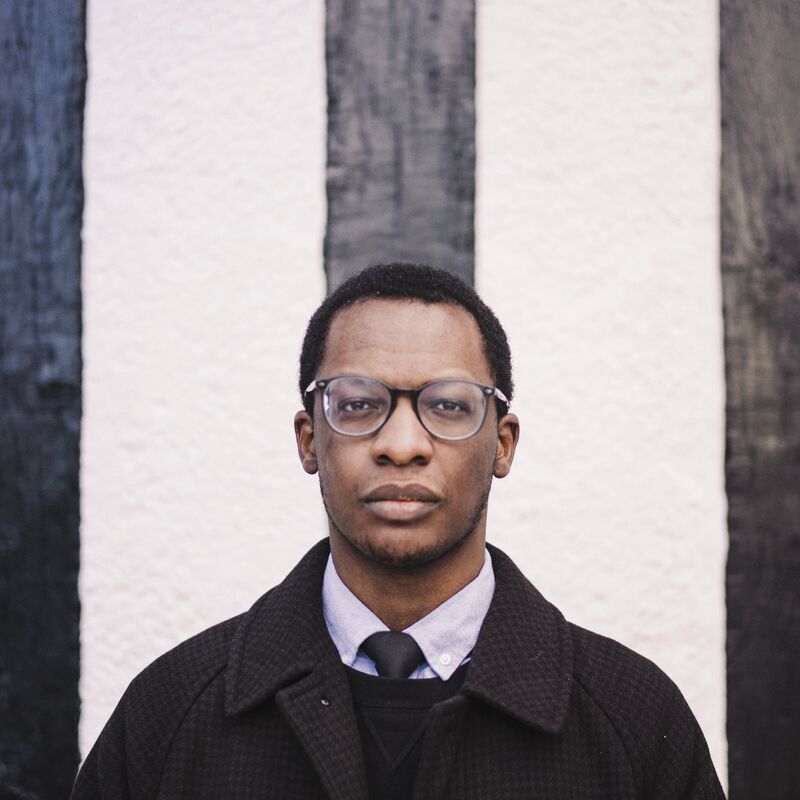 Author Tomiwa Owolade stands in front of a door wearing a black coat, shirt and tie