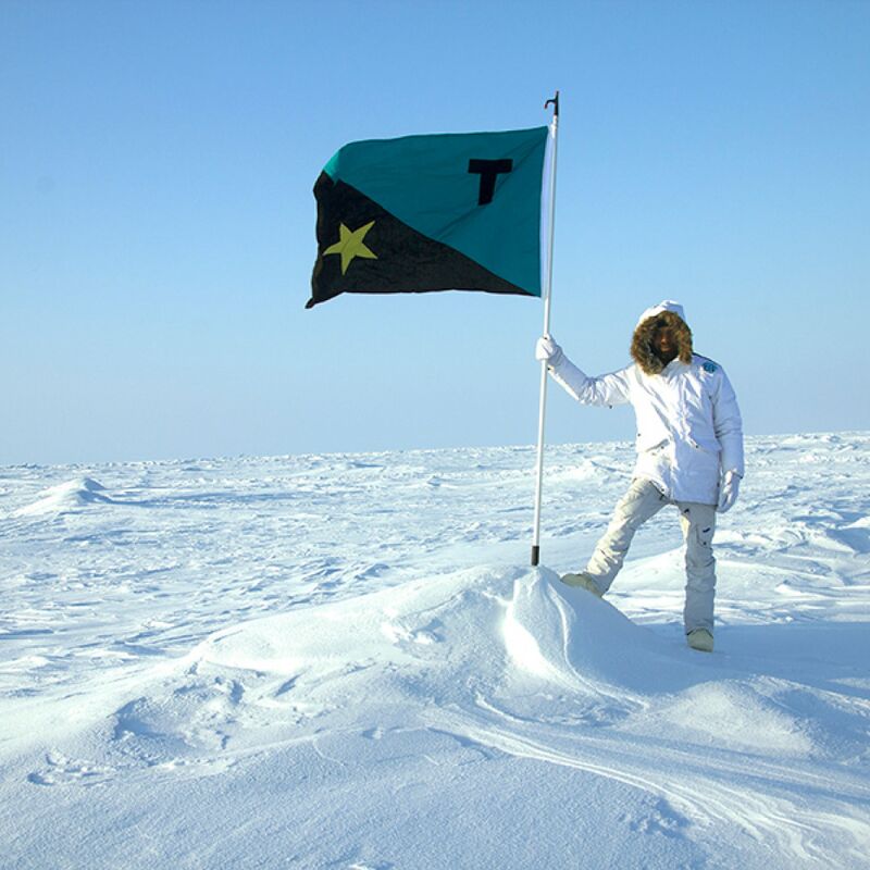 Artist Tavares Strachan, a Black man in his 30s, stands at the North Pole on a white landscape of ice and snow, wearing an all white snowsuit. He is holding a flag pole, on top of which is a flag that is aquamarine and black divided diagonally with a Gold star on the lower black half and a black 't' on the upper aquamarine half