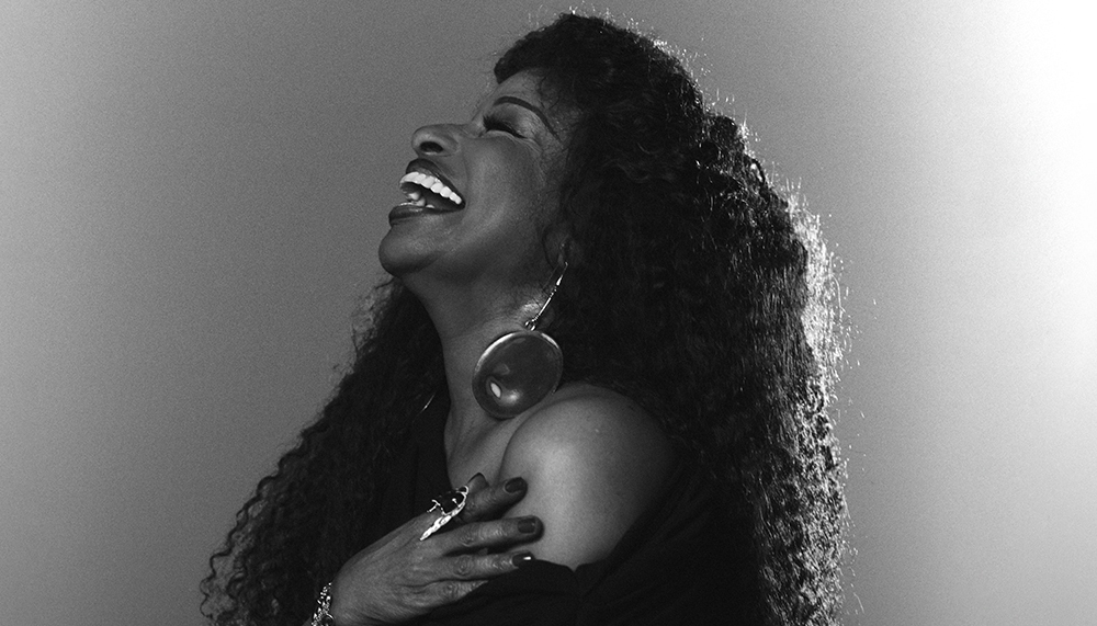 Black and white side profile of the singer Chaka Khan, laughing. She is wearing an over the shoulder black dress.