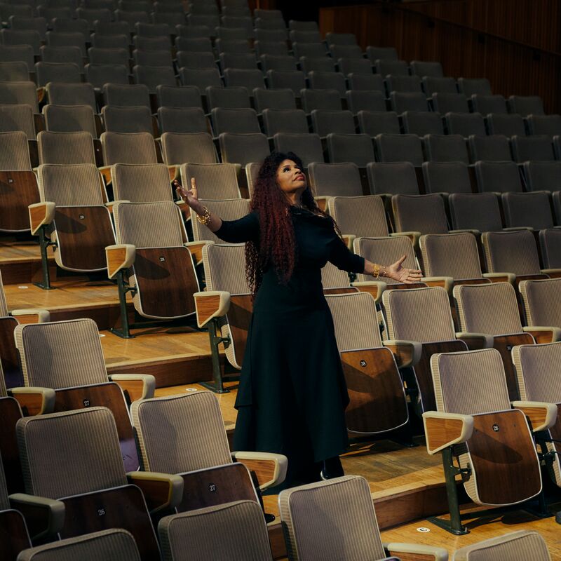 The musician Chaka Khan, a Black woman with long curly hair wears a long black dress and stands with her arms stretched outwards among the seats of the Royal Festival Hall auditorium