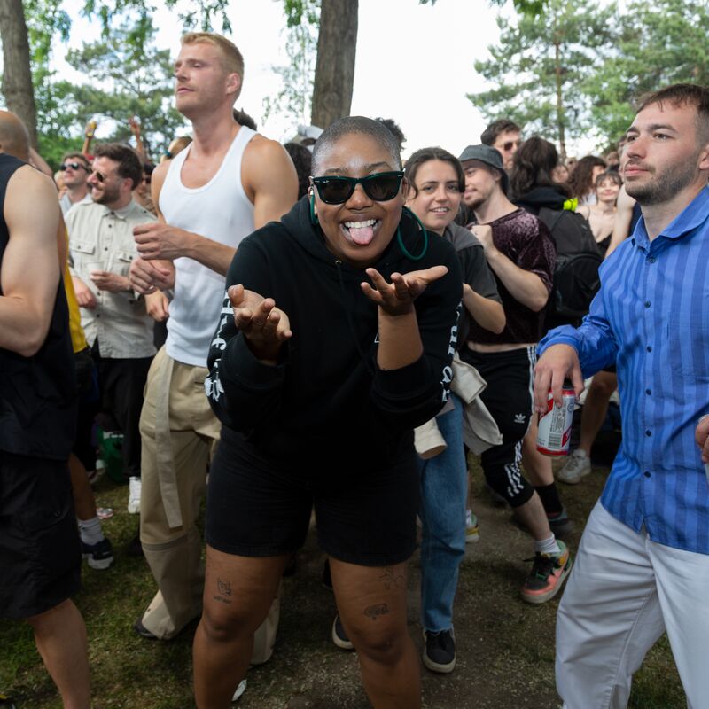 People dancing outdoors at a London Queer Soundsystem event. Person wearing black sunglasses, shorts and jumper looks at the camera with their tongue out