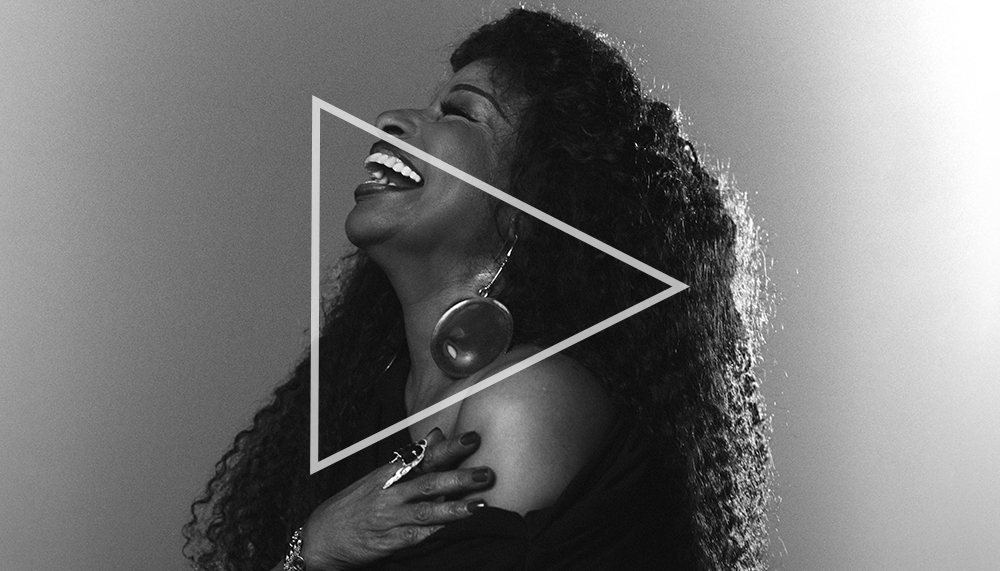 Black and white side profile of the singer Chaka Khan, laughing. She is wearing an over the shoulder black dress.