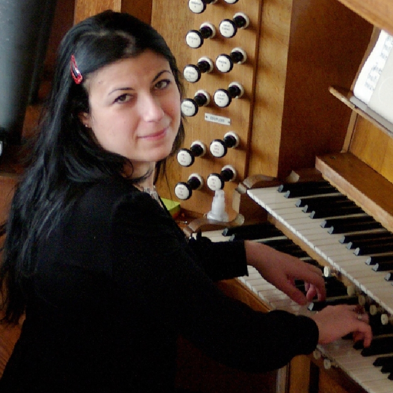 Ourania Gassiou sits with their hads on the keys of an organ as they look over their shoulder at the camera. 