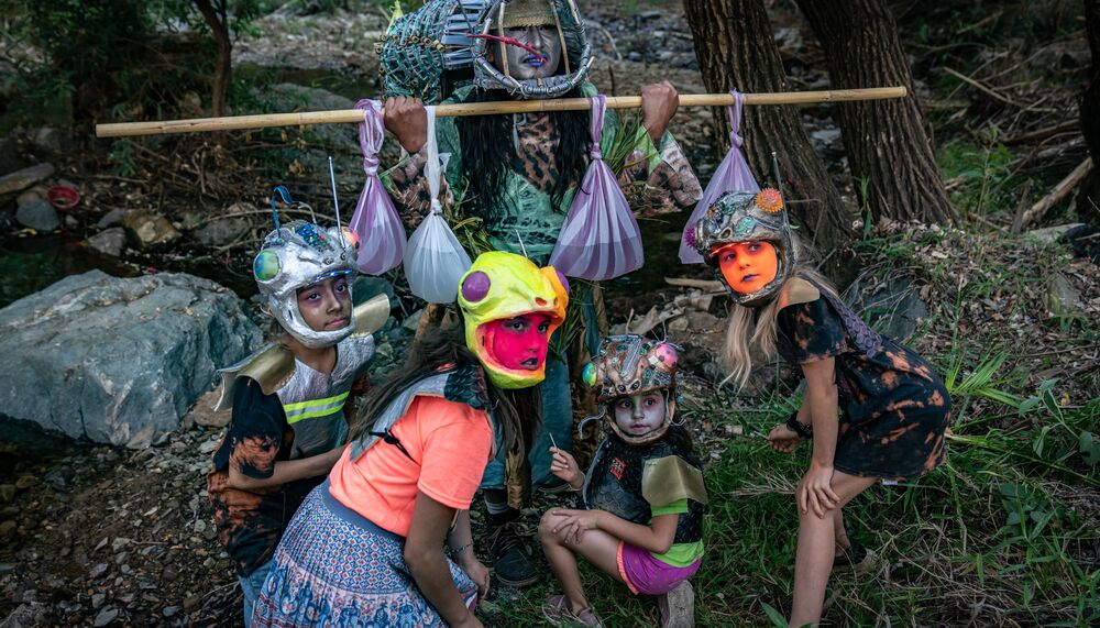 A women in a mask holding a wooden pole with plastic bags hanging off. There are 4 children in helmets crouching in front.