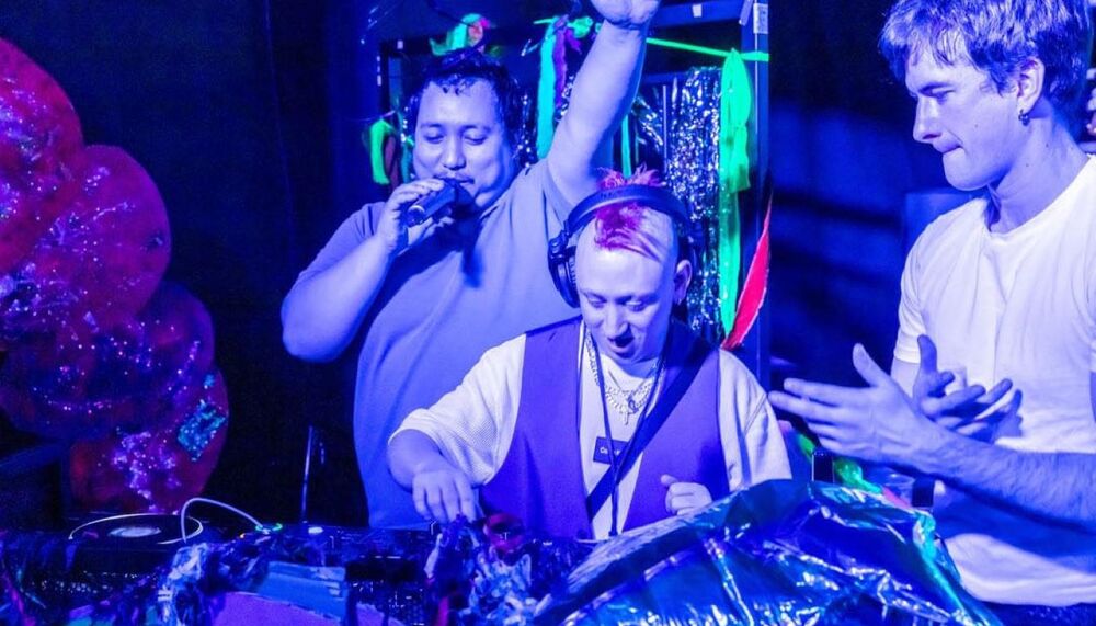 Three people stand around DJ decks lit by blue and purple lighting.