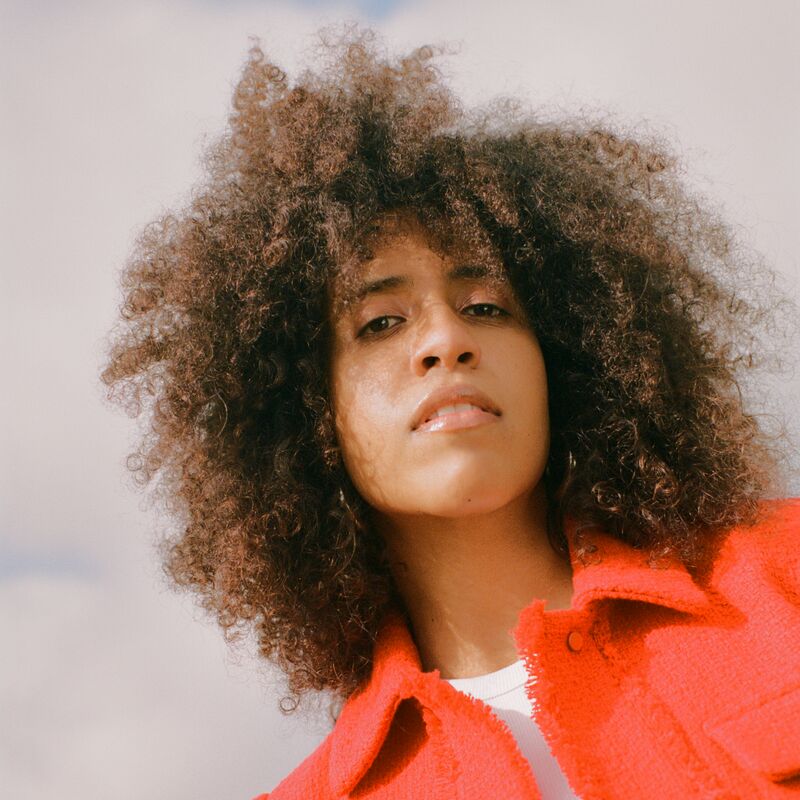 Singer and DJ Naima Adams wearing an orange shirt looks down at the camera with the sky behind her.