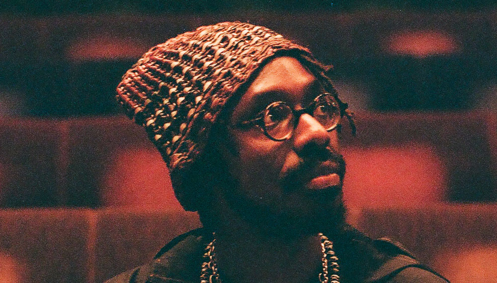 Artist SHABAKA sits in an empty auditorium and gazes off camera.