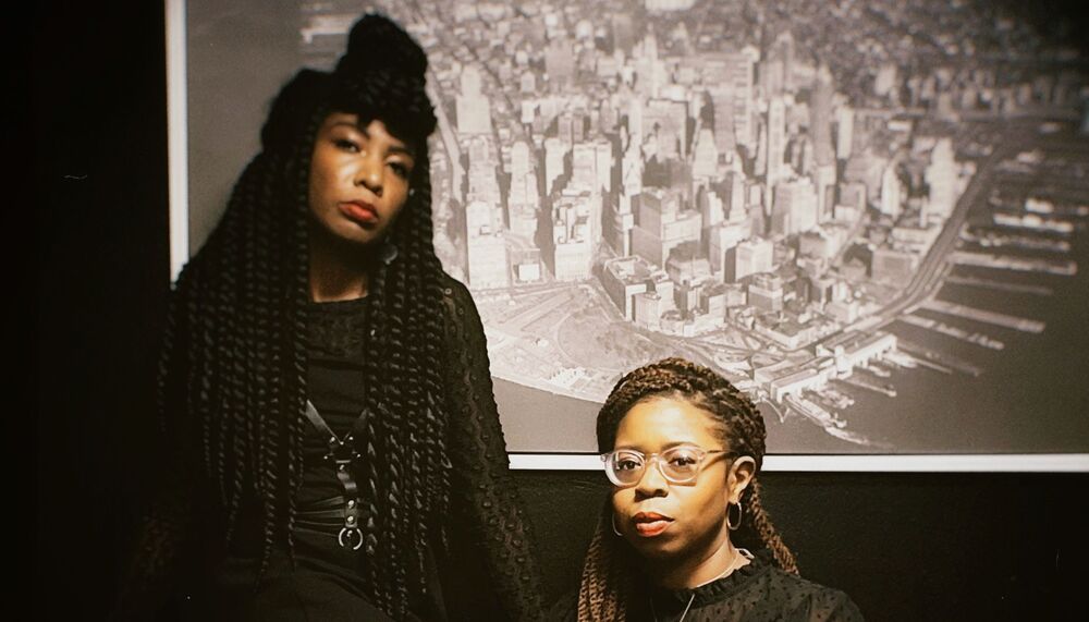 Members of Big Joanie sit on a red leather sofa wearing black dresses with a black and white photo of a city behind them.