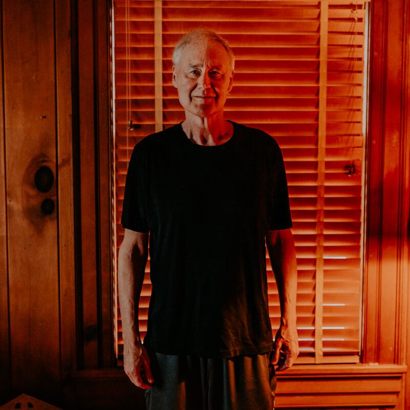 Musician Bruce Hornsby stands in front of a wooden wall with a window covered by blinds, wearing a black T-Shirt.