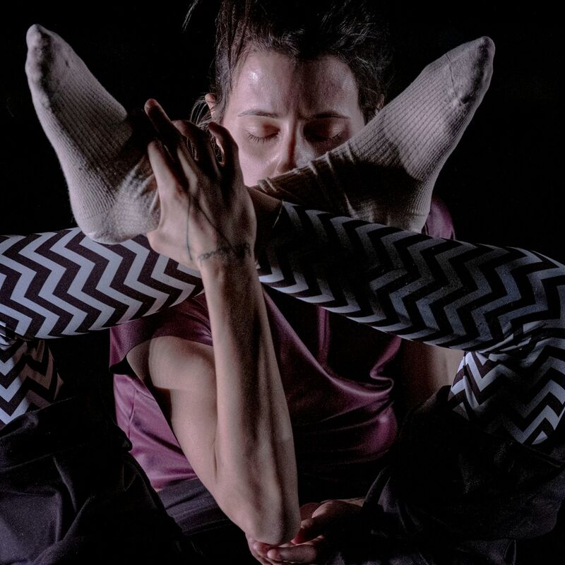 Dancer in contorted position, holding their feet by their head, against a black background. They wear a black and white zigzagged leggings and a pink shirt.