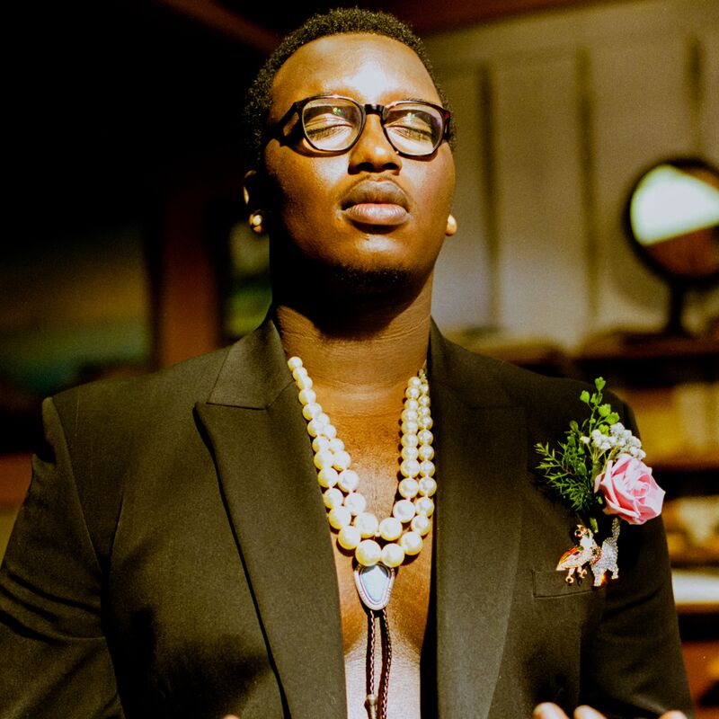 Artist SIPHO. wearing a black blazer with a bare chest and necklaces, holding their palms up in front of them and their eyes closed.