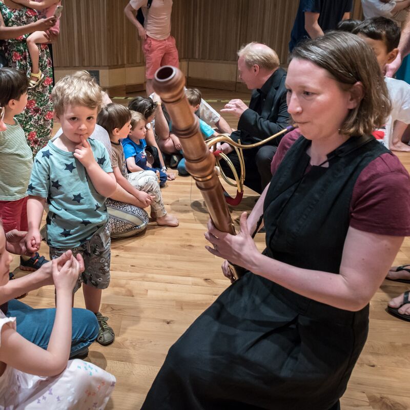 A woman with short, brown hair playing the Bassoon in front of toddlers and their carers