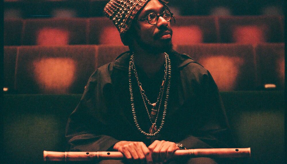 Artist Shabaka sits in an empty auditorium holding a wooden flute in his hands as he gazes off camera.