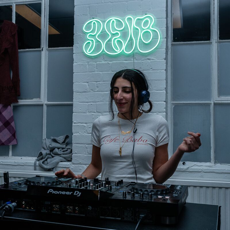 A woman stands with headphones on in front of DJ decks.