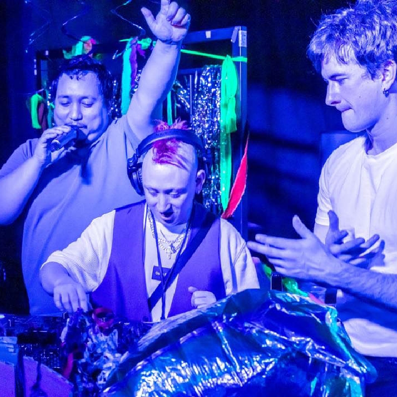 Three people stand around DJ decks lit by blue and purple lighting.