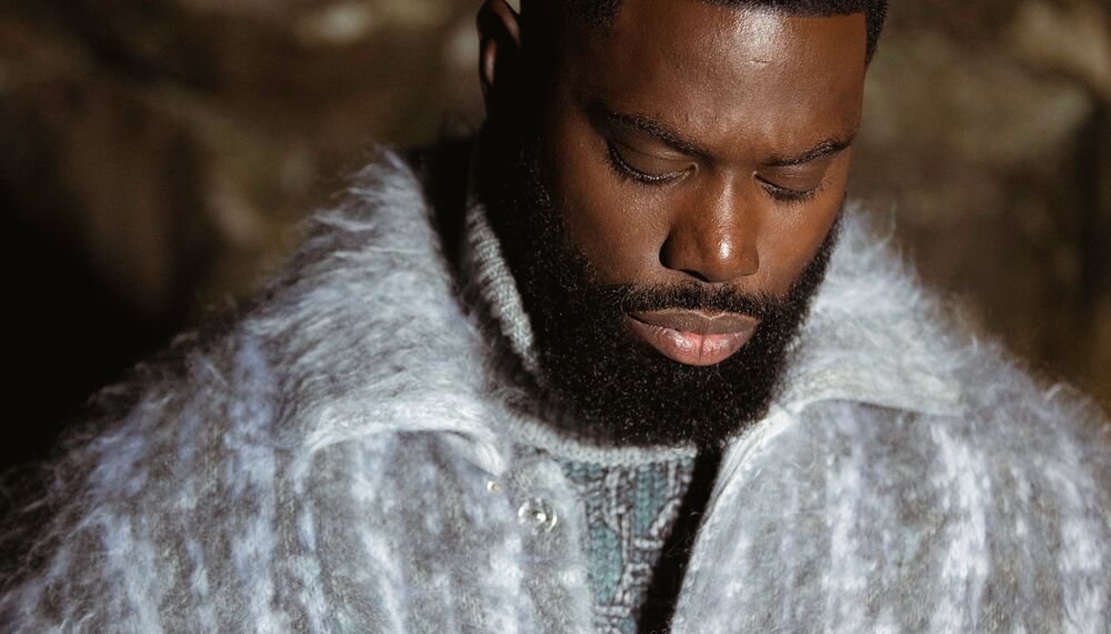 Ghetts stands against a rocky background wearing a light blue jacket.