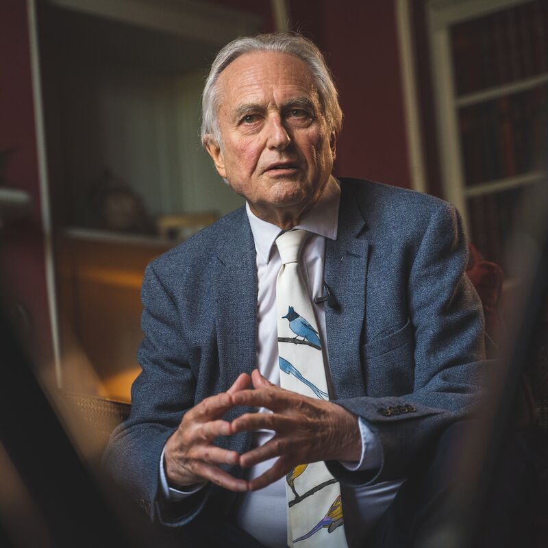 Richard Dawkins wears a blue suit sitting in front of a bookshelf