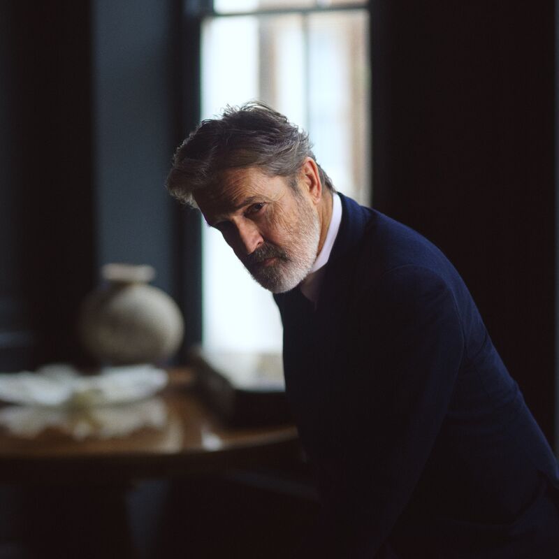 Rupert Everett sits in a dark room, leaning forward and wearing a navy blazer.