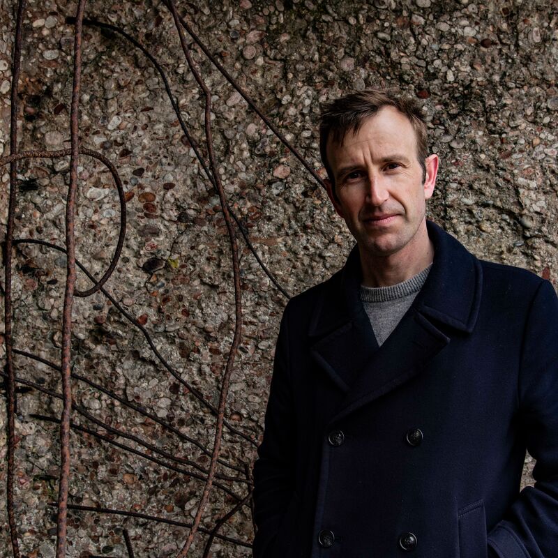 Robert Macfarlane stands against stone background wearing a navy blue coat