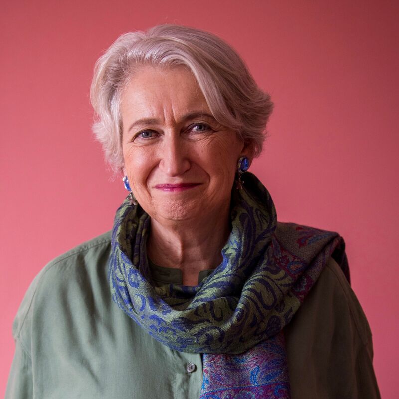 Lindsey Hilsum stands against a pink background wearing a green jumper and scarf.