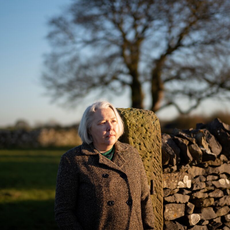 Woman in a brown jacket stands by a wall in a field.