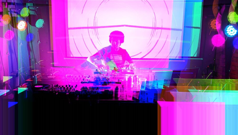 A DJ stands at decks with blue, green and pink lights bouncing round the image.