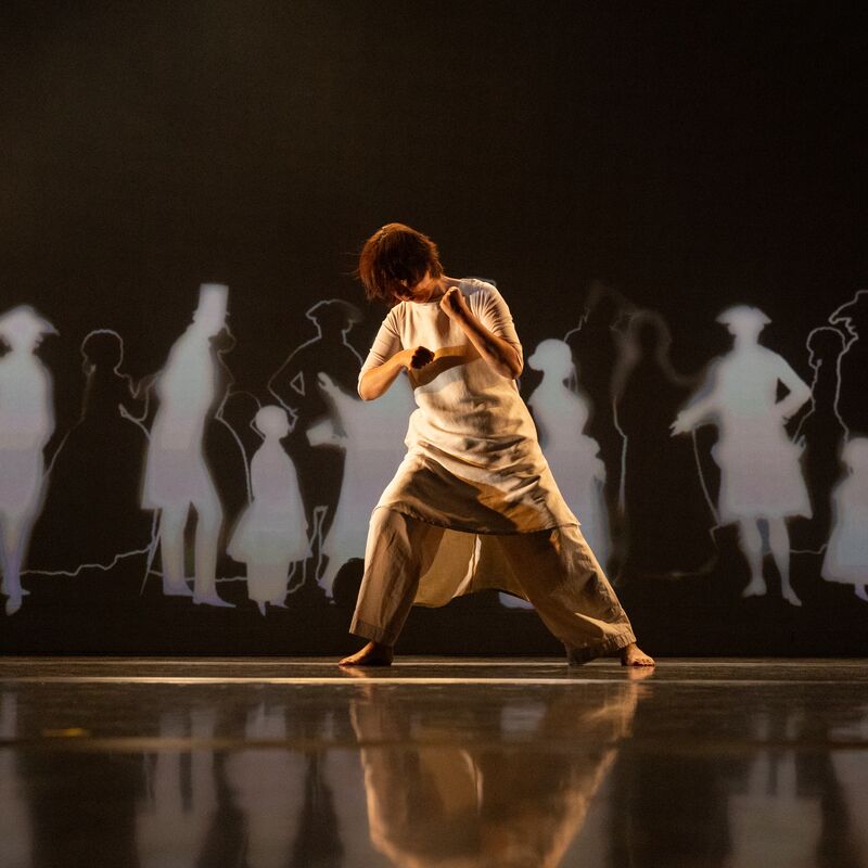 Chisato Minamimura dancing on stage wearing a loose-fitting cream tunic and trousers. Black and white silhouettes of figures in historical costume are projected on the wall behind her.