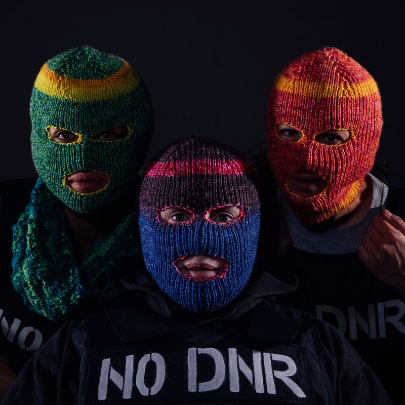 Three people stare directly at the camera and are each wearing different coloured, knitted balaclavas where only their eyes and mouths are exposed. They are all wearing black stab vests with the words ‘NO DNR’ stencilled onto the vests in white paint.