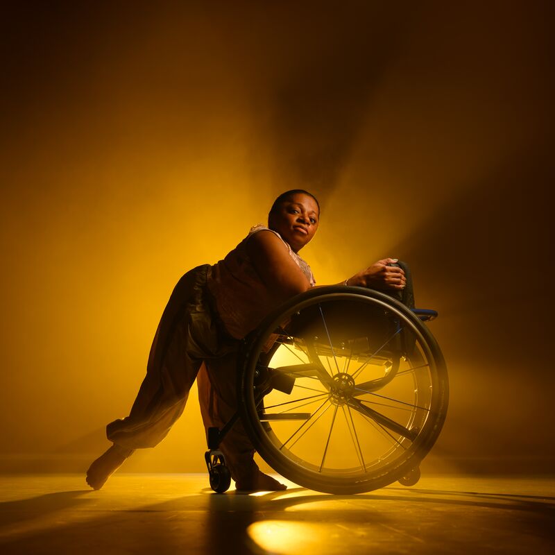 A black female dancer is leaning against her wheelchair facing towards it, in front of an amber light