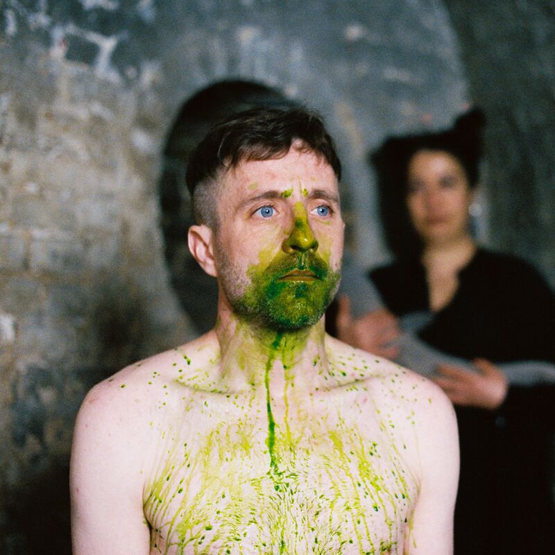 A shirtless person with green liquid all over their face and down their chest. Behind them is anther person wearing black and brick walls.