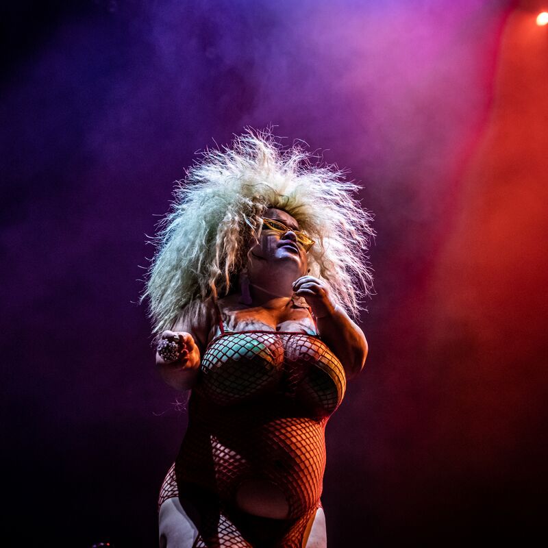 Midgitte Bardot stands in a fishnet dress and green bra with bright blonde hair that fans out against the purple and red lit backdrop.