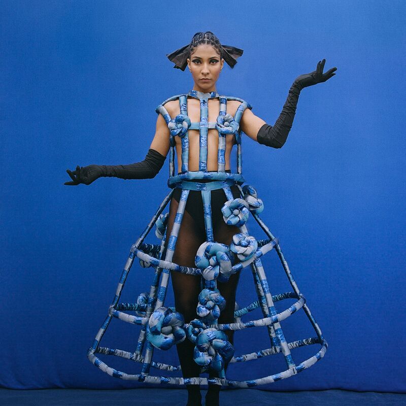 A woman poses in a blue cage dress standing against a blue background.