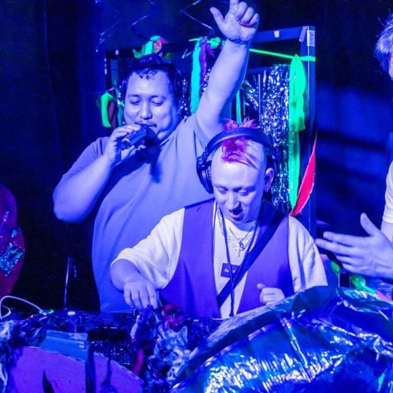 Three people stand around DJ decks lit by blue and purple lighting