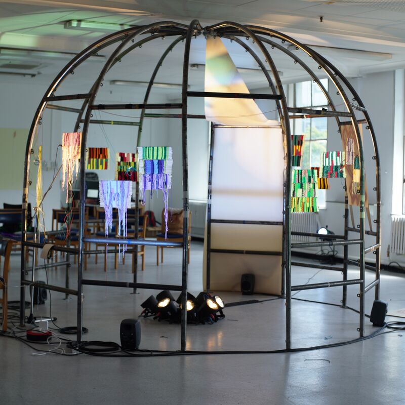 A domed steel structure sits in the centre of a room. Weaved art hangs from the poles