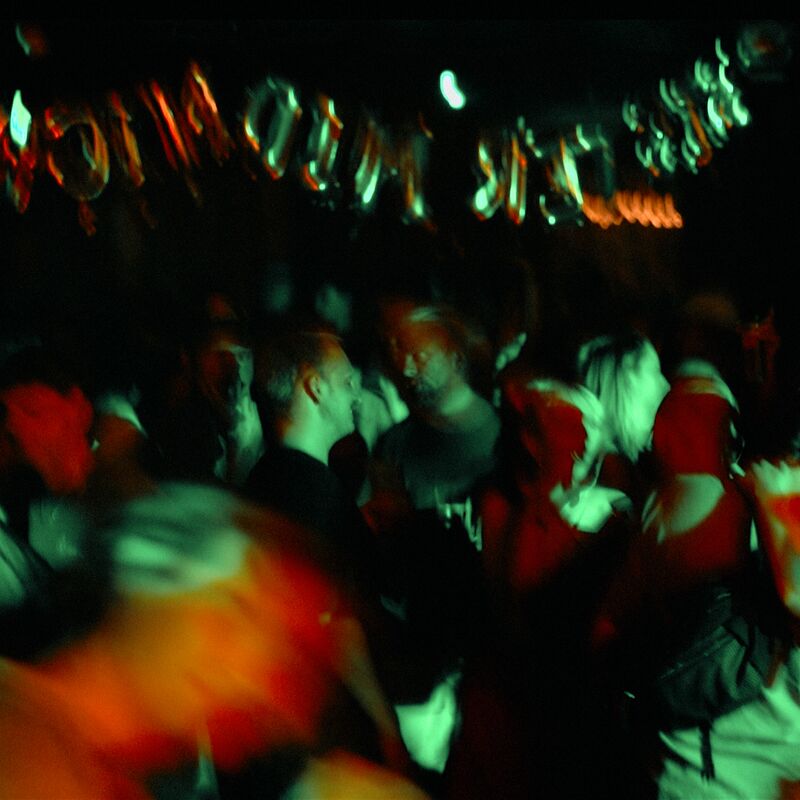 A crowd of people dancing in a dark venue under red and green flights. Silver letter shaped balloons hang above them