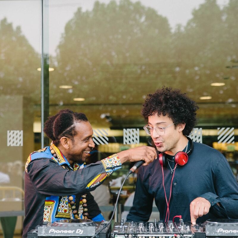 A DJ stands in front of a set of decks while a performer holds a microphone to his mouth