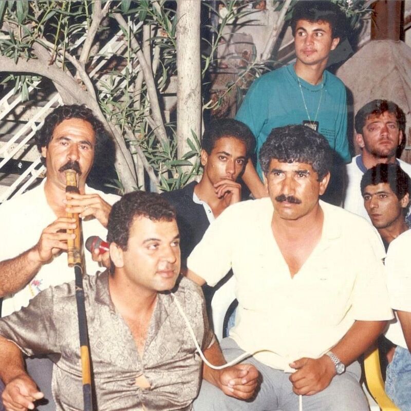 A group of men sat together, one plays a wooden floot while another holds a microphone