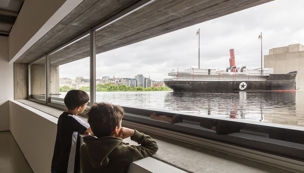 An installation view of kids watching a boat floating in black water with a brutalist building in the background.