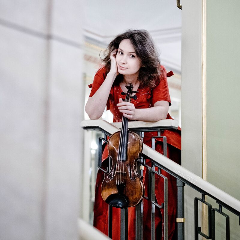 Woman stands on a staircase holding a violin, she is wearing a red dress.