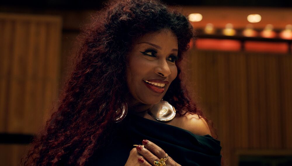 Head shot of Chaka Khan in the Royal Festival Hall, looking to the side and smiling with red-tinted curly hair and large gold earrings.